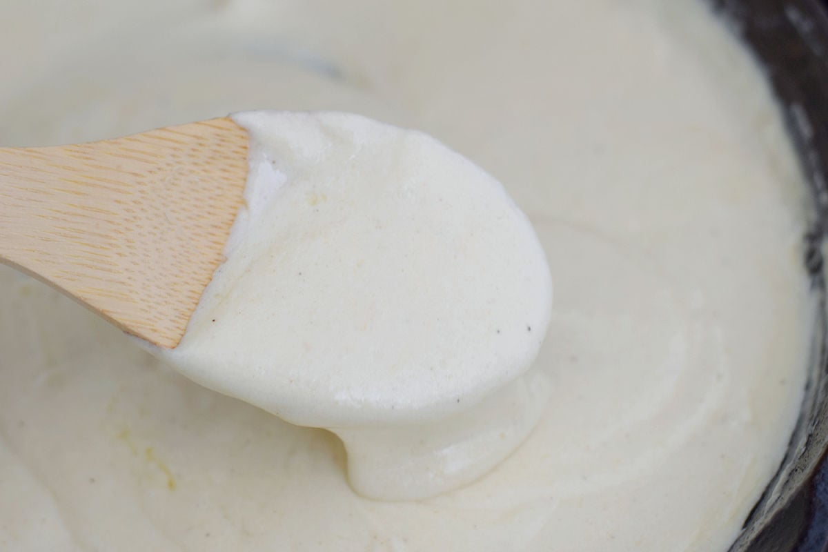 Close-up of Alfredo sauce coating a wooden spoon above a skillet of sauce.
