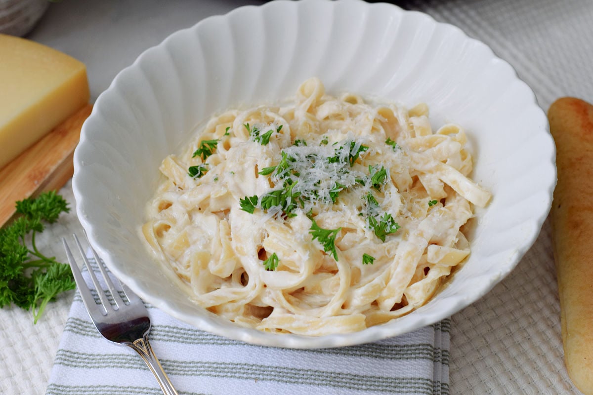 Fettuccine coated in Alfredo sauce and topped with grated cheese and parsley in a white scalloped bowl.