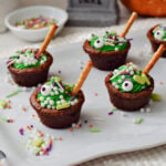 Finished cauldron brownie bites with green frosting, candy eyeballs, white sprinkle bubbles, and pretzel-stick spoons.