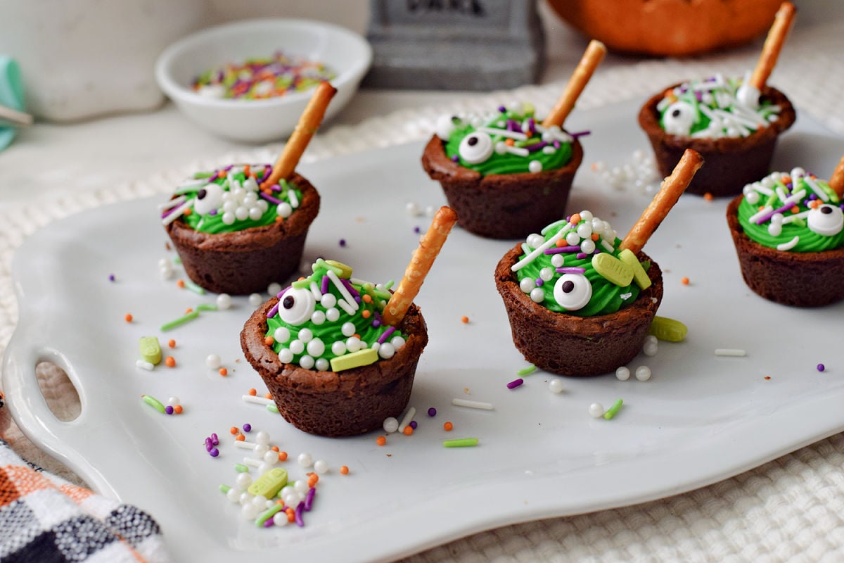Finished cauldron brownie bites with green frosting, candy eyeballs, white sprinkle bubbles, and pretzel-stick spoons.