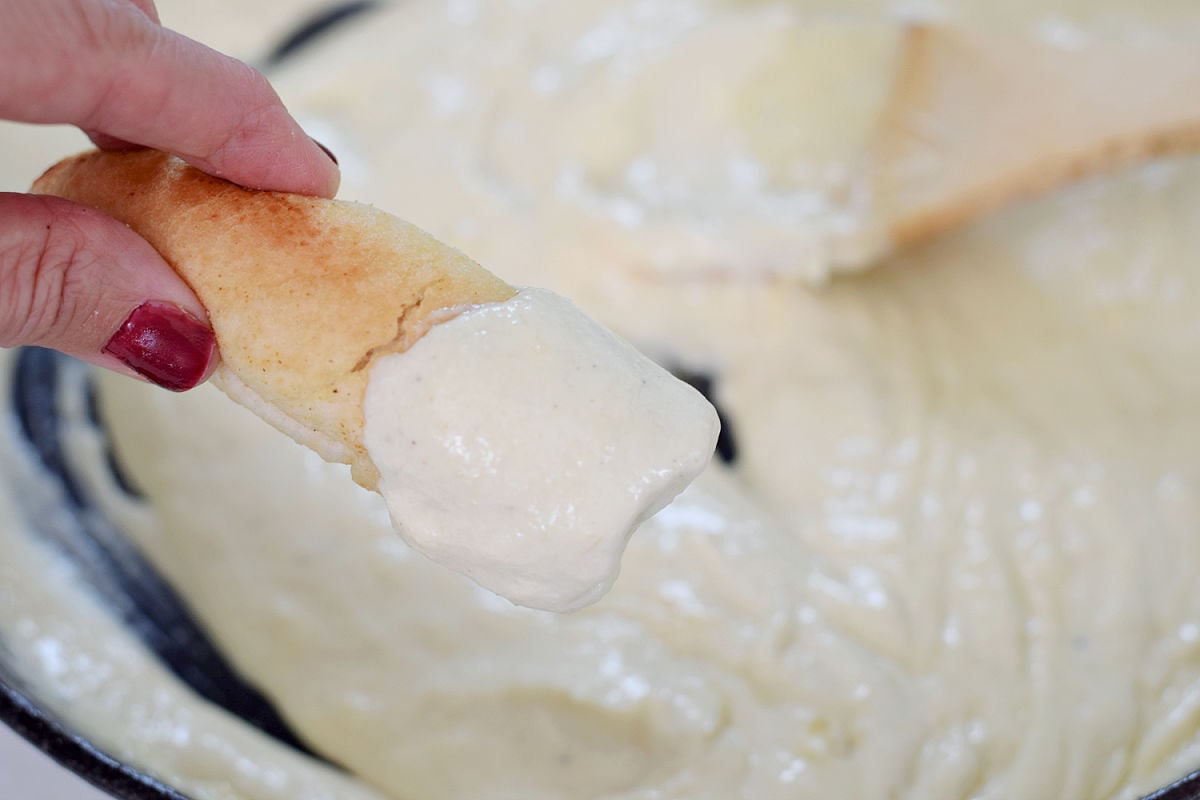 Hand holding a breadstick coated in creamy Alfredo sauce above a cast iron skillet.