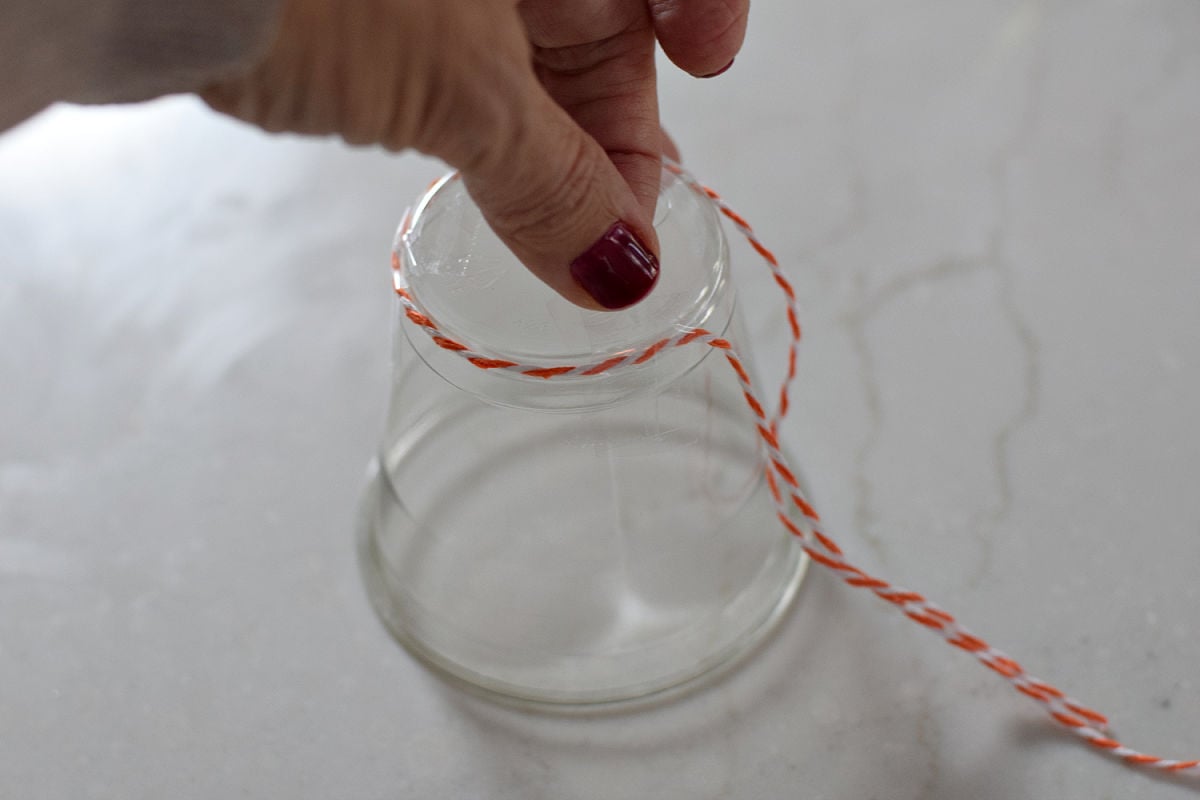 Hand pressing tape over orange-and-white twine wrapped around the narrow end of an upside-down clear plastic cup.