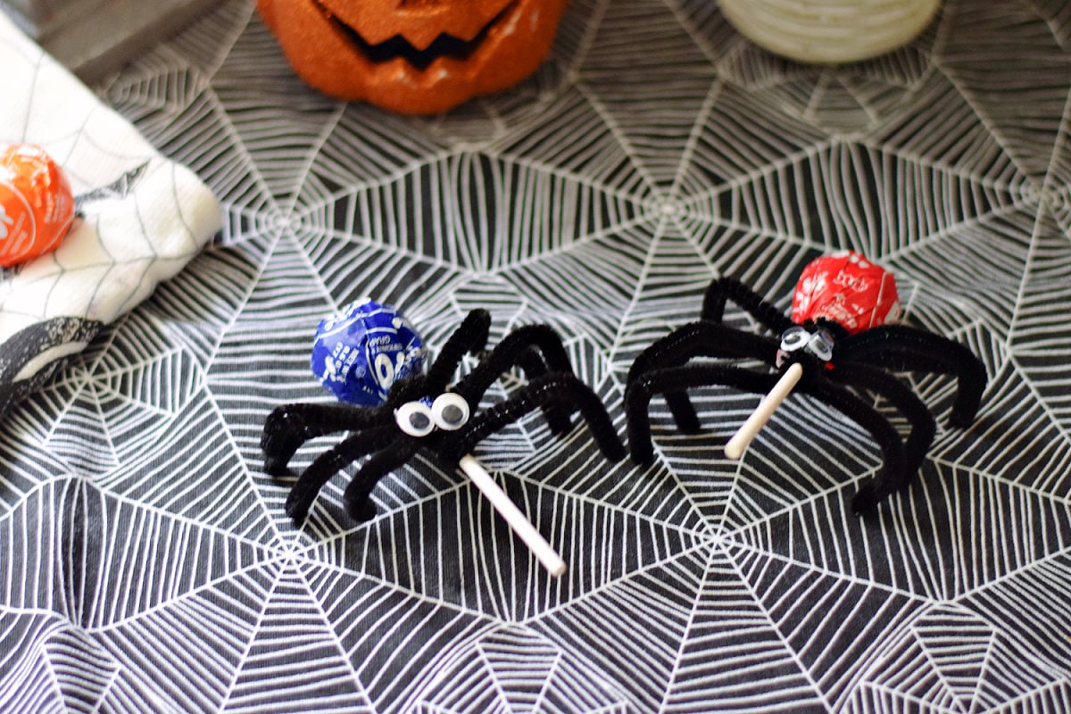 Lollipop spiders made with wrapped Tootsie Pops, black pipe cleaner legs, and googly eyes on spiderweb fabric.