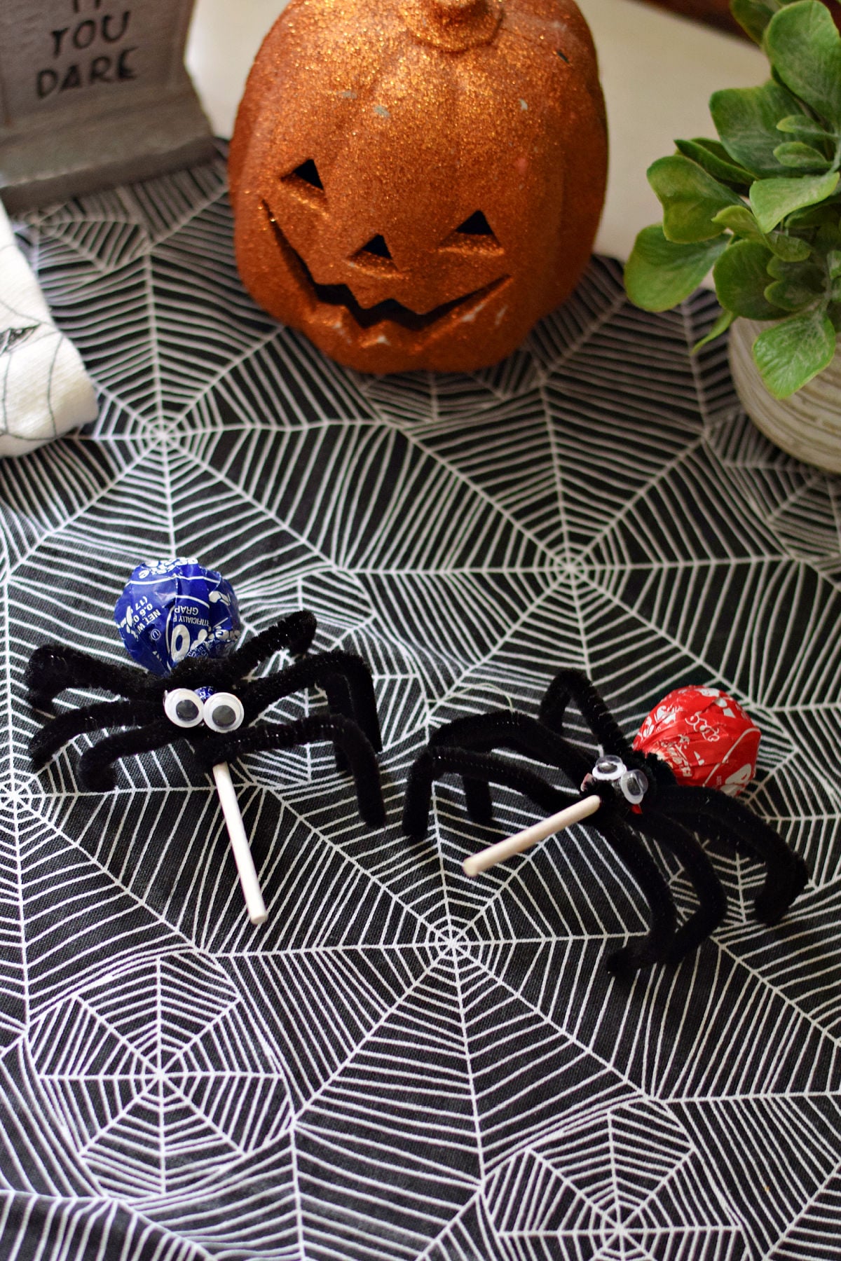 Finished Tootsie Pop spiders on spiderweb fabric with a jack-o’-lantern decoration behind them.
