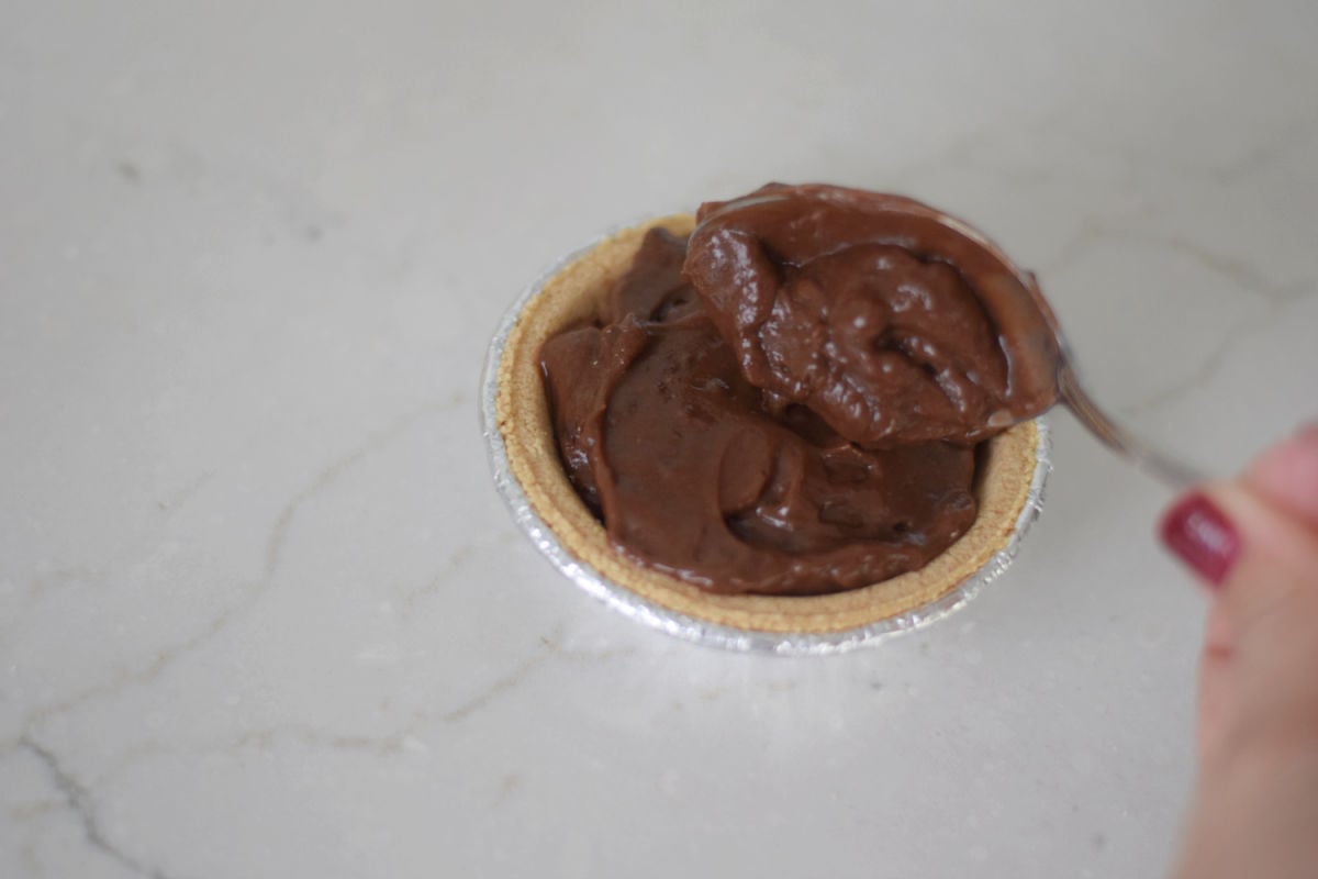 Spoon adding chocolate pudding to a mini graham cracker pie crust in a foil tin.
