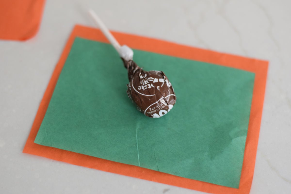 A wrapped Tootsie Pop centered on green tissue paper layered over a slightly larger orange rectangle.