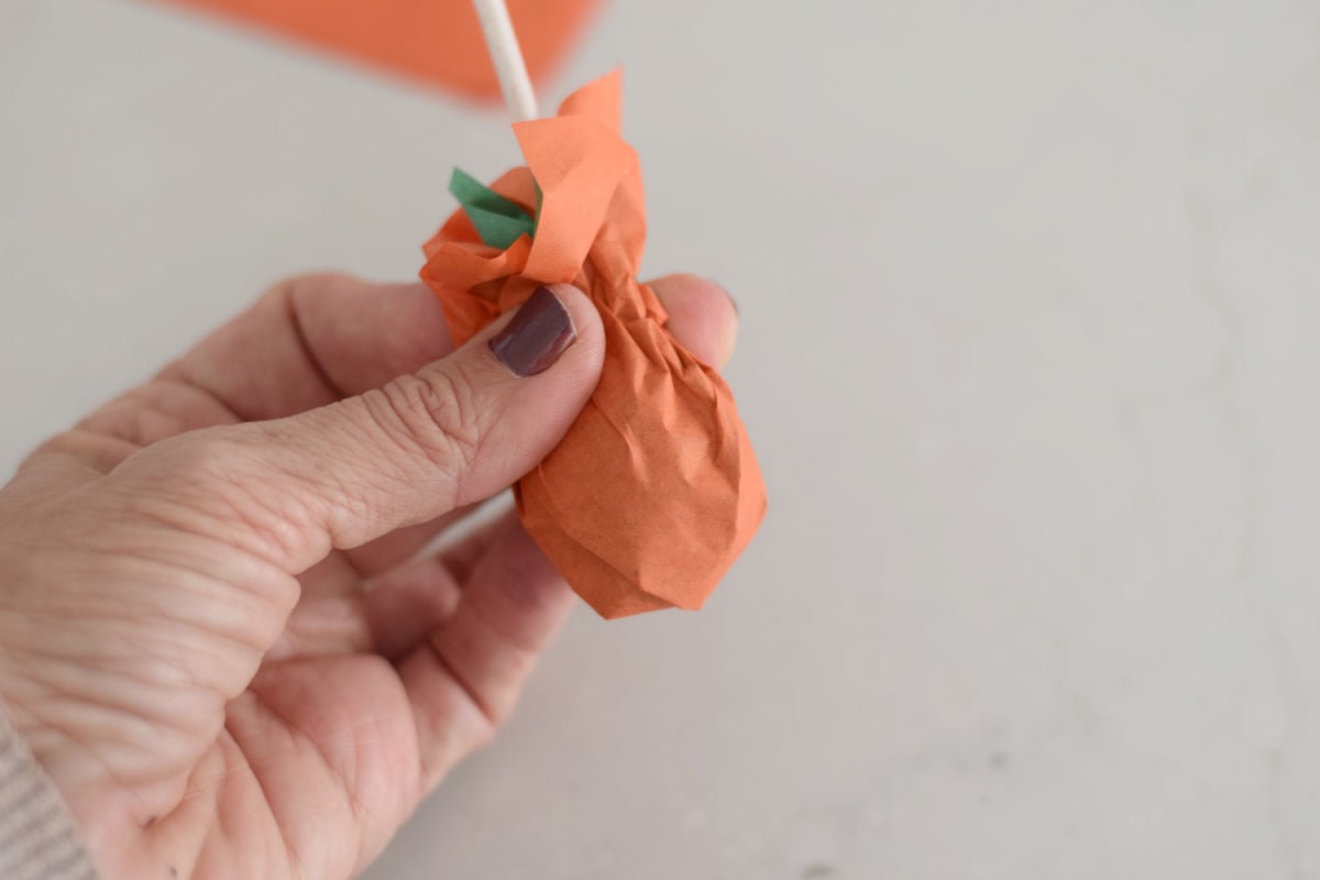  Hand gathering orange and green tissue paper around a lollipop with the stick pointing upward.