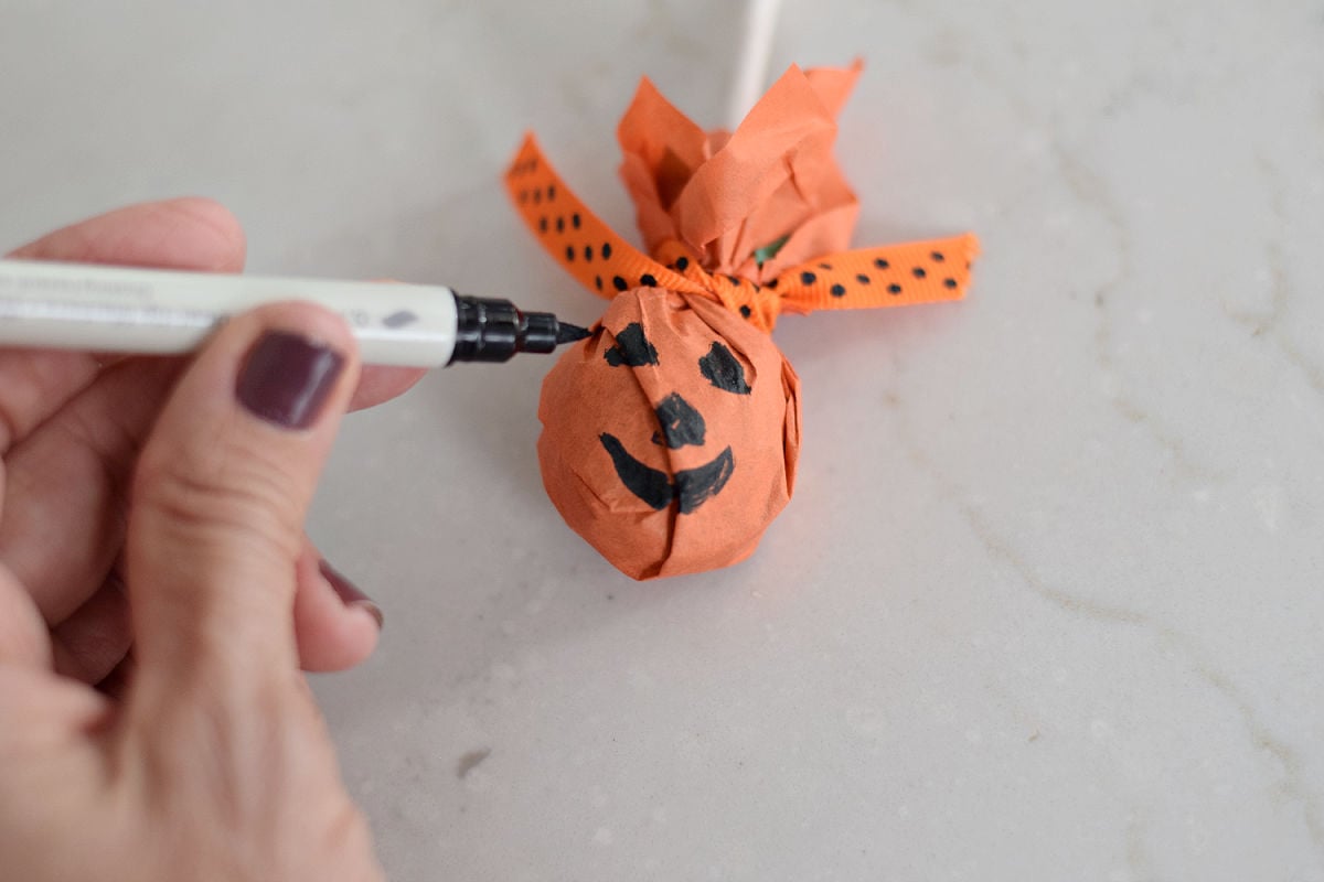 Drawing a black jack-o’-lantern face on an orange tissue-paper lollipop tied with polka-dot ribbon.