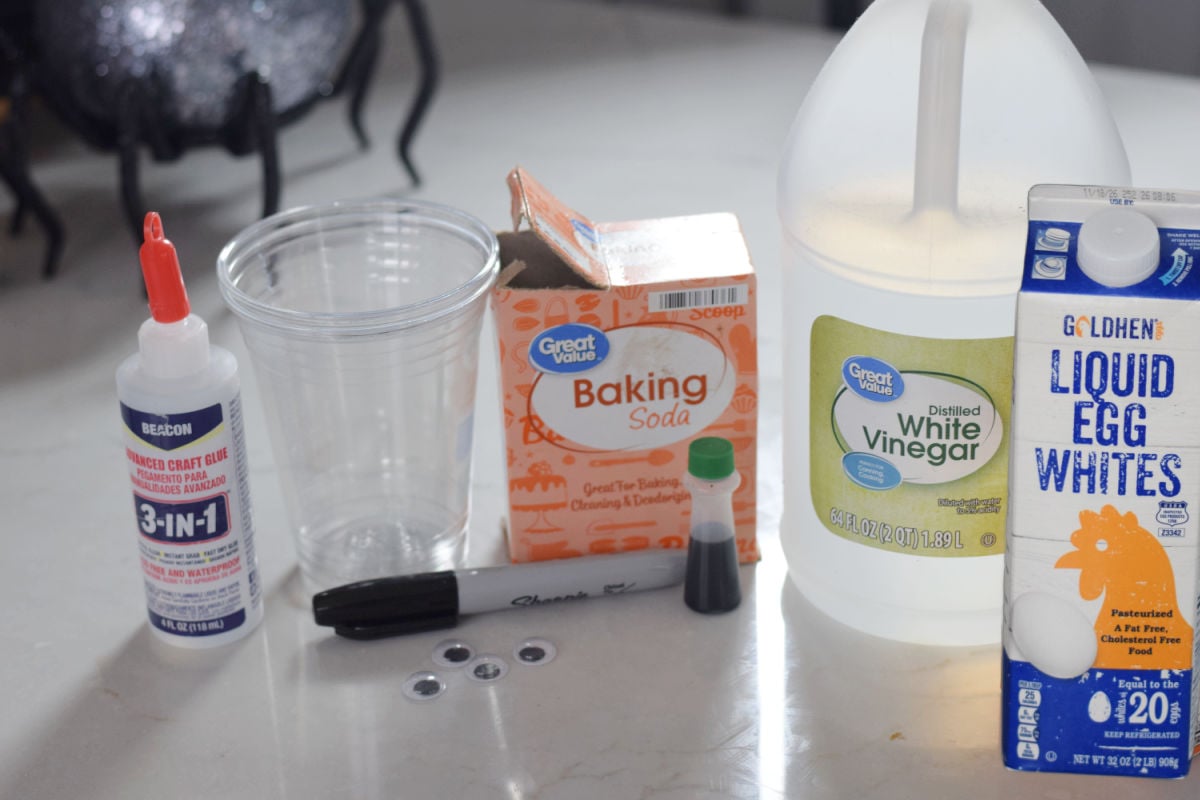 Supplies for a Frankenstein foam science experiment, including a clear plastic cup, baking soda, liquid egg whites, white vinegar, green food coloring, black marker, googly eyes, and craft glue.