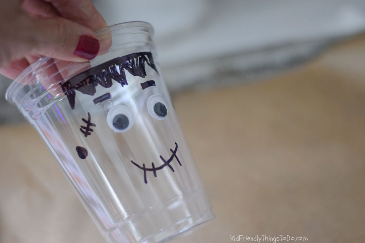 Hand holding a clear plastic cup decorated with black Frankenstein hair, a stitched smile, and googly eyes.