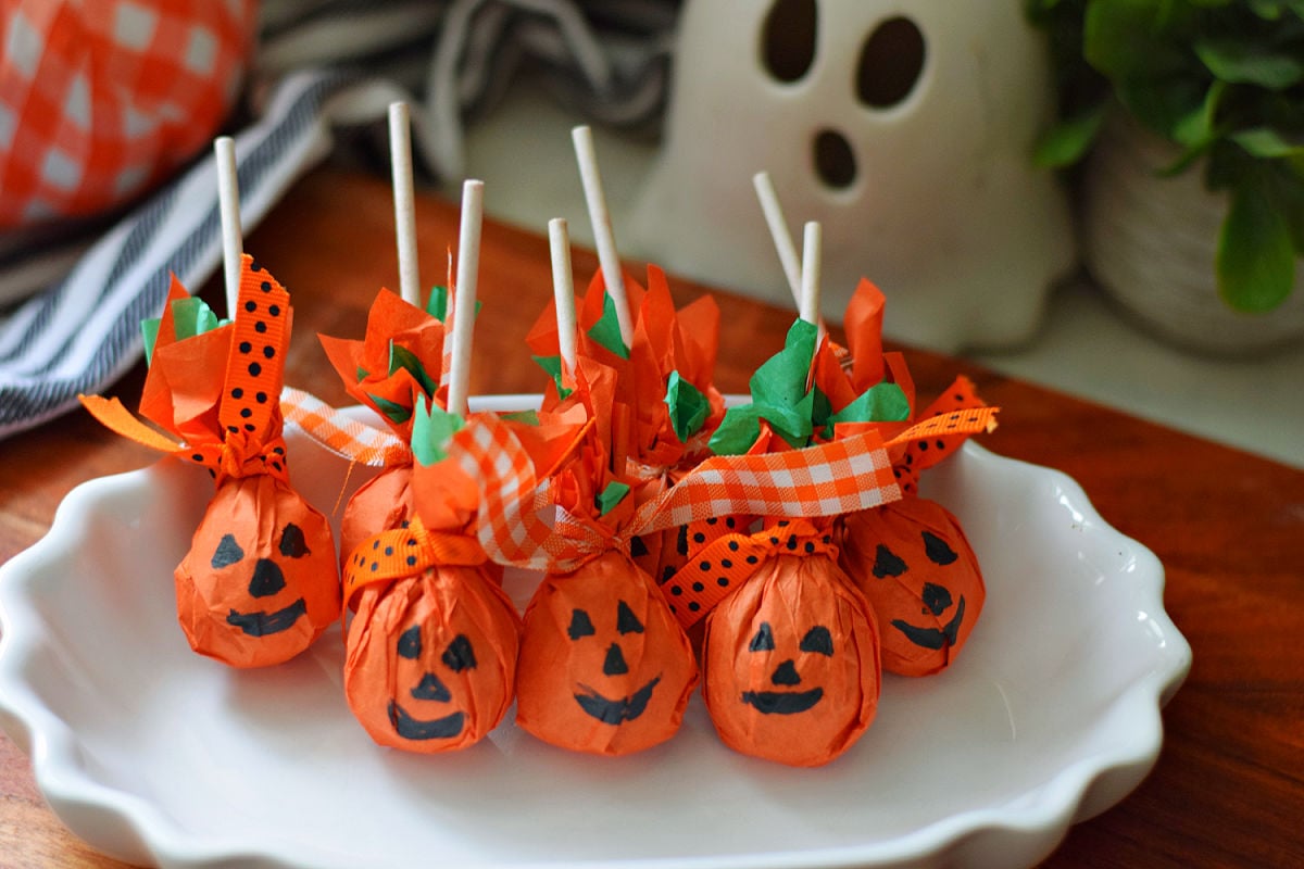 Pumpkin lollipops wrapped in orange tissue paper with green tissue accents, ribbon ties, and black jack-o’-lantern faces on a white platter.