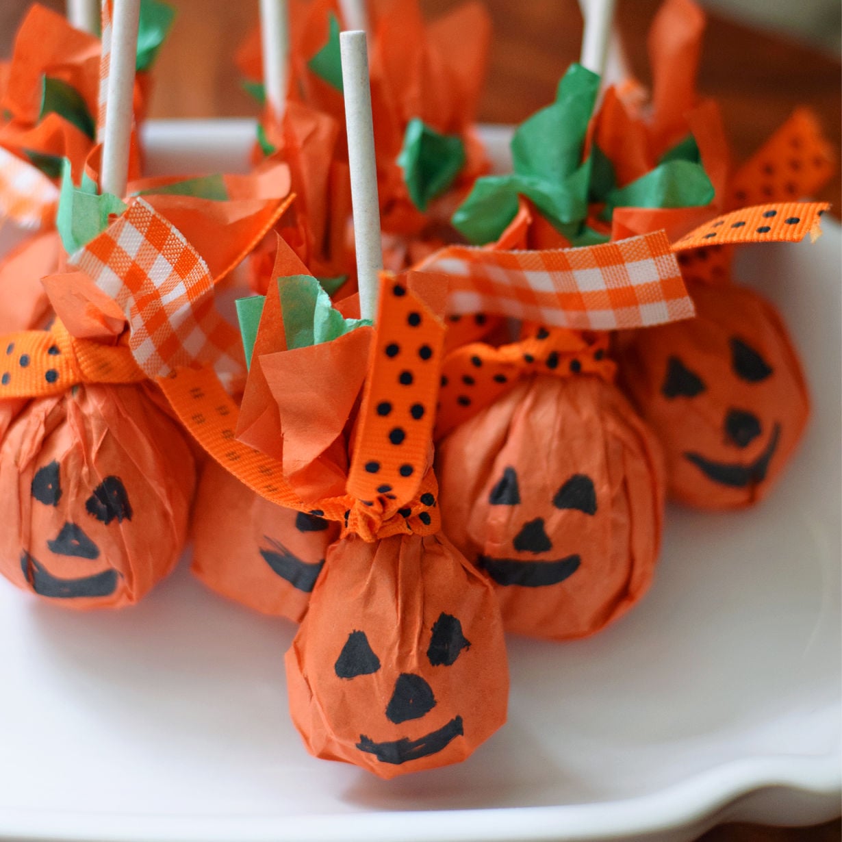 Close-up of tissue-paper jack-o’-lantern lollipops with orange ribbons and small green accents on a white platter.