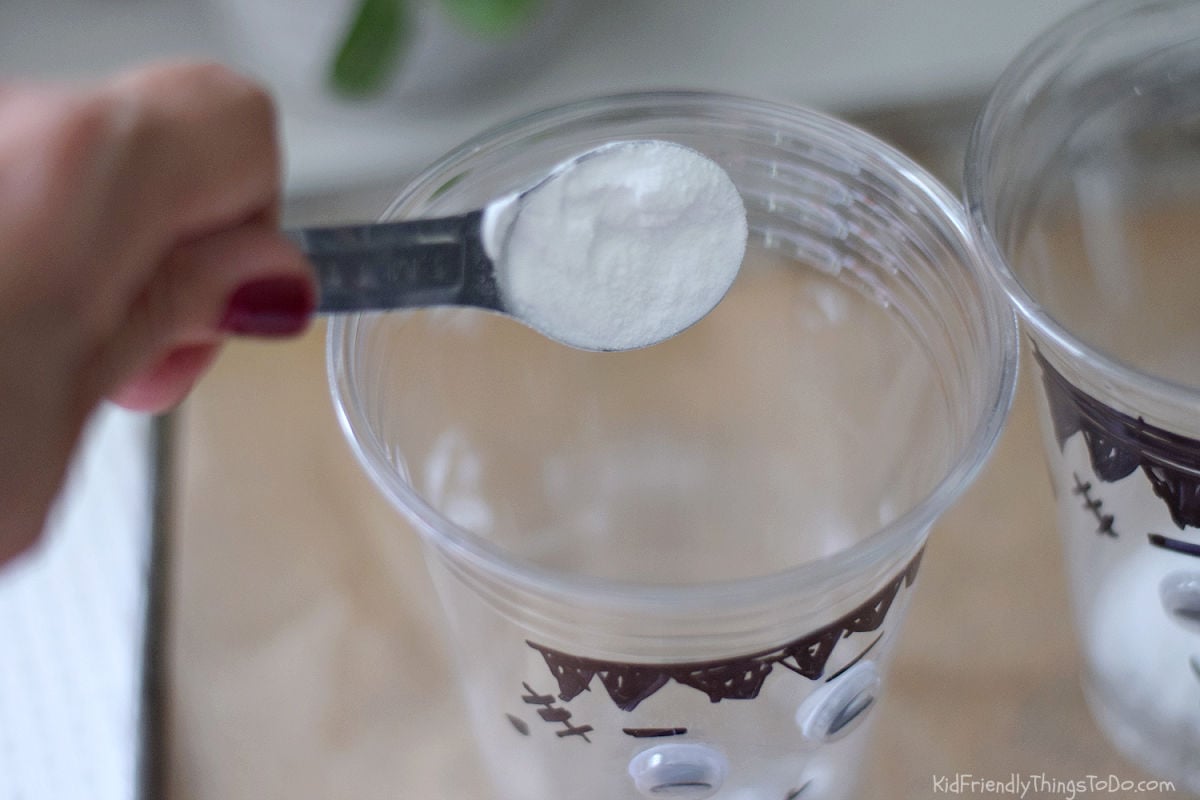 Adding a spoonful of baking soda to a clear cup decorated with a Frankenstein face.