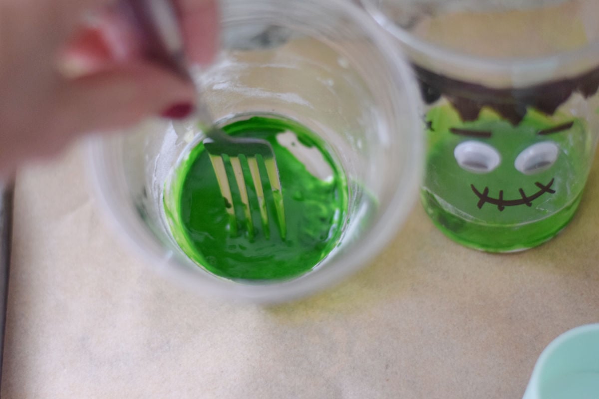 Stirring baking soda, liquid egg whites, and green food coloring with a fork inside a Frankenstein cup.