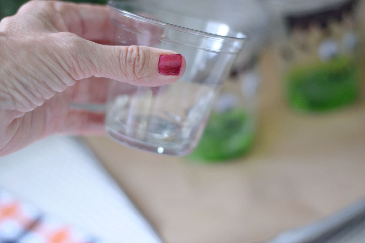 Hand holding a small clear cup of measured vinegar beside a prepared Frankenstein cup.