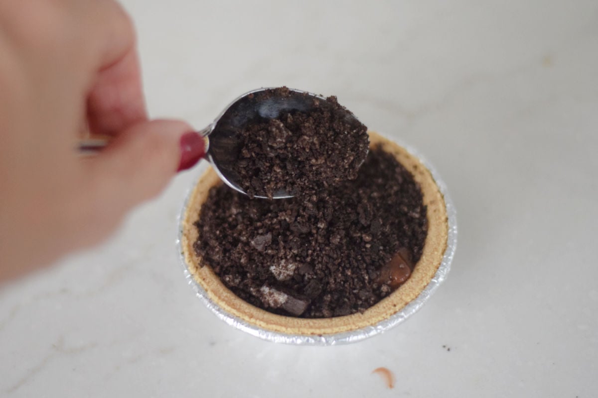 Spoon spreading crushed Oreo cookies over chocolate pudding in a mini graham cracker pie crust.
