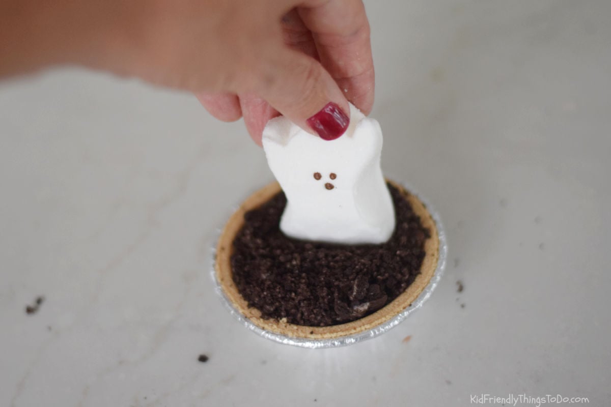 Hand placing a white ghost Peep upright on Oreo crumbs covering a mini chocolate pudding pie.