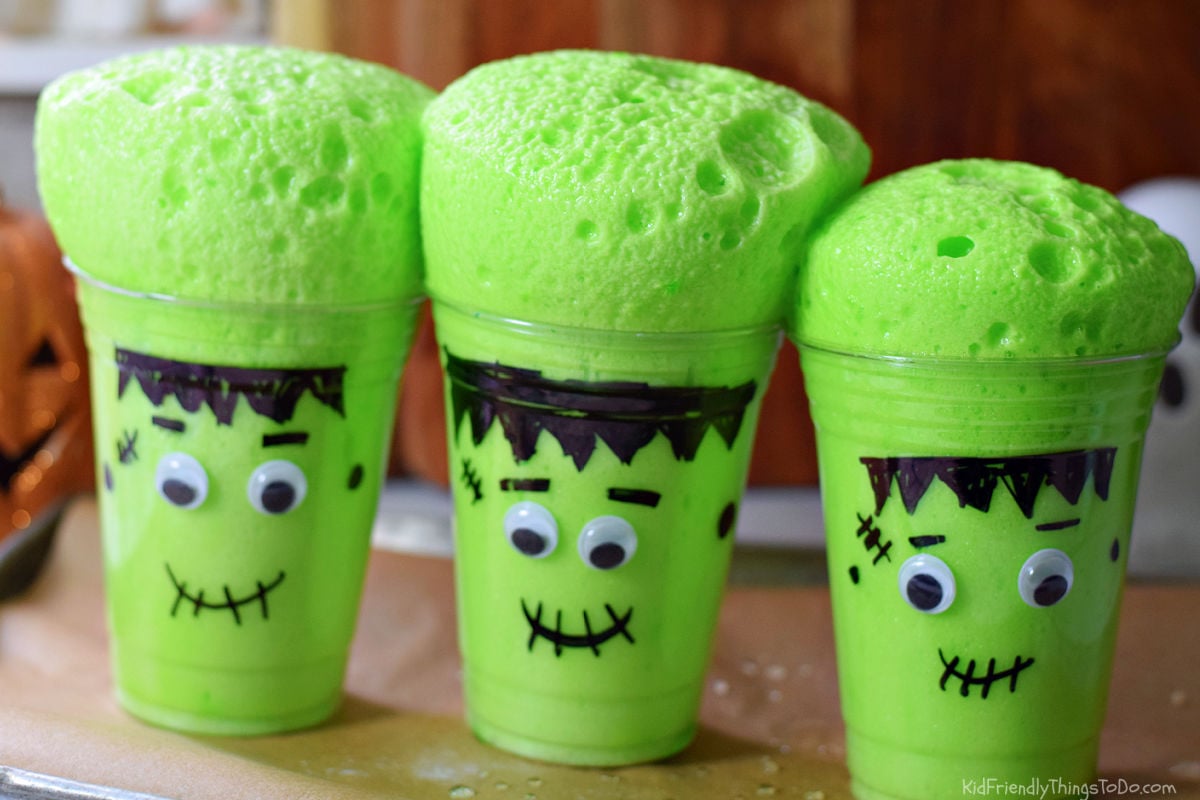 Three clear cups decorated with Frankenstein faces topped with tall crowns of bubbly green foam.