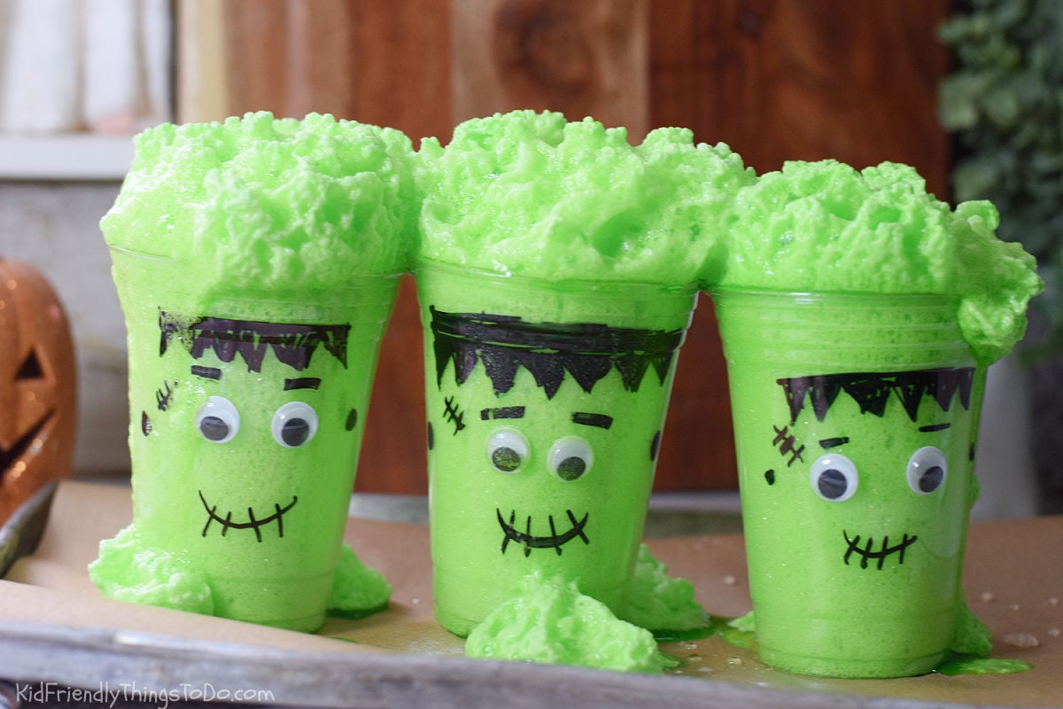 Green foam spilling over the rims and down the sides of three Frankenstein cups onto a baking tray.