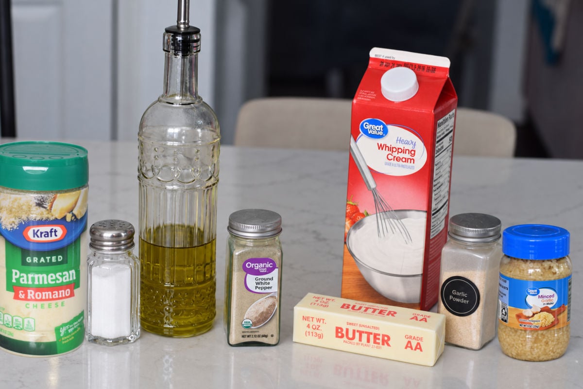Unsalted butter, olive oil, heavy cream, minced garlic, garlic powder, white pepper, salt, and grated Parmesan and Romano cheese arranged on a kitchen counter.