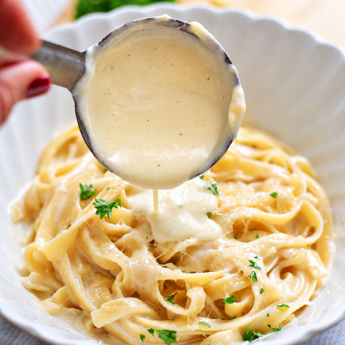 Alfredo sauce pouring from a metal ladle onto fettuccine topped with grated cheese and parsley in a white bowl.
