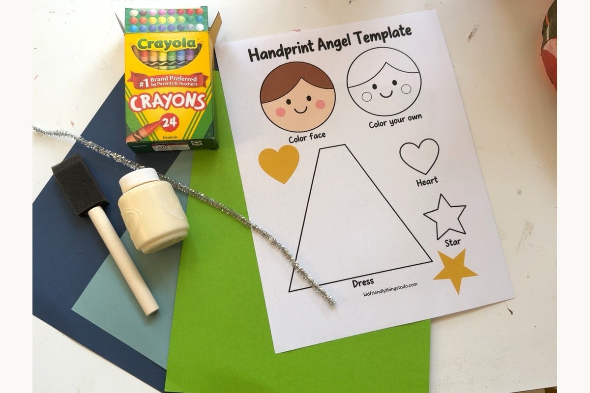Angel handprint craft supplies with printable template, colored paper, crayons, paint, foam brush, and a silver pipe cleaner