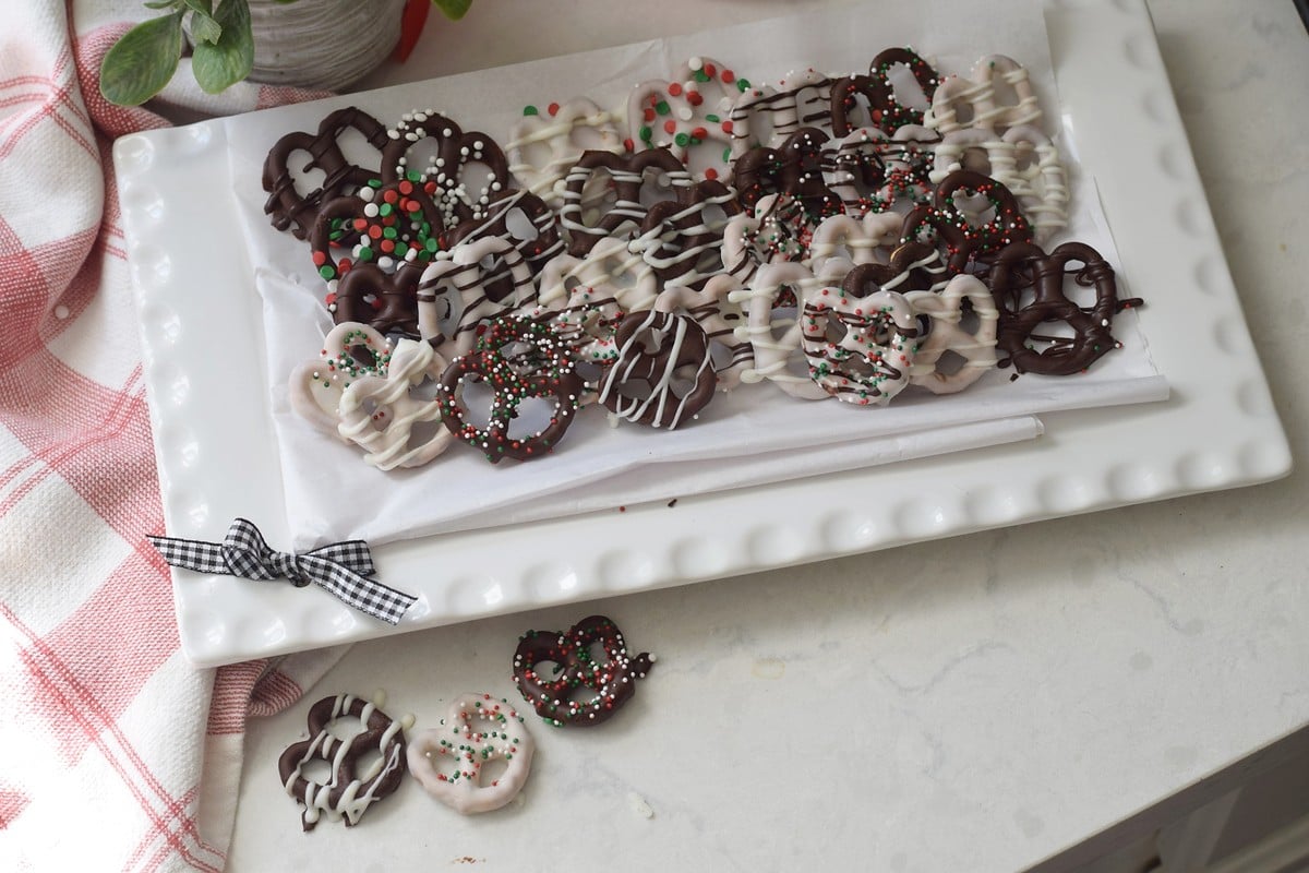 Chocolate covered pretzels with white drizzle and red, green, and white sprinkles on a white platter.