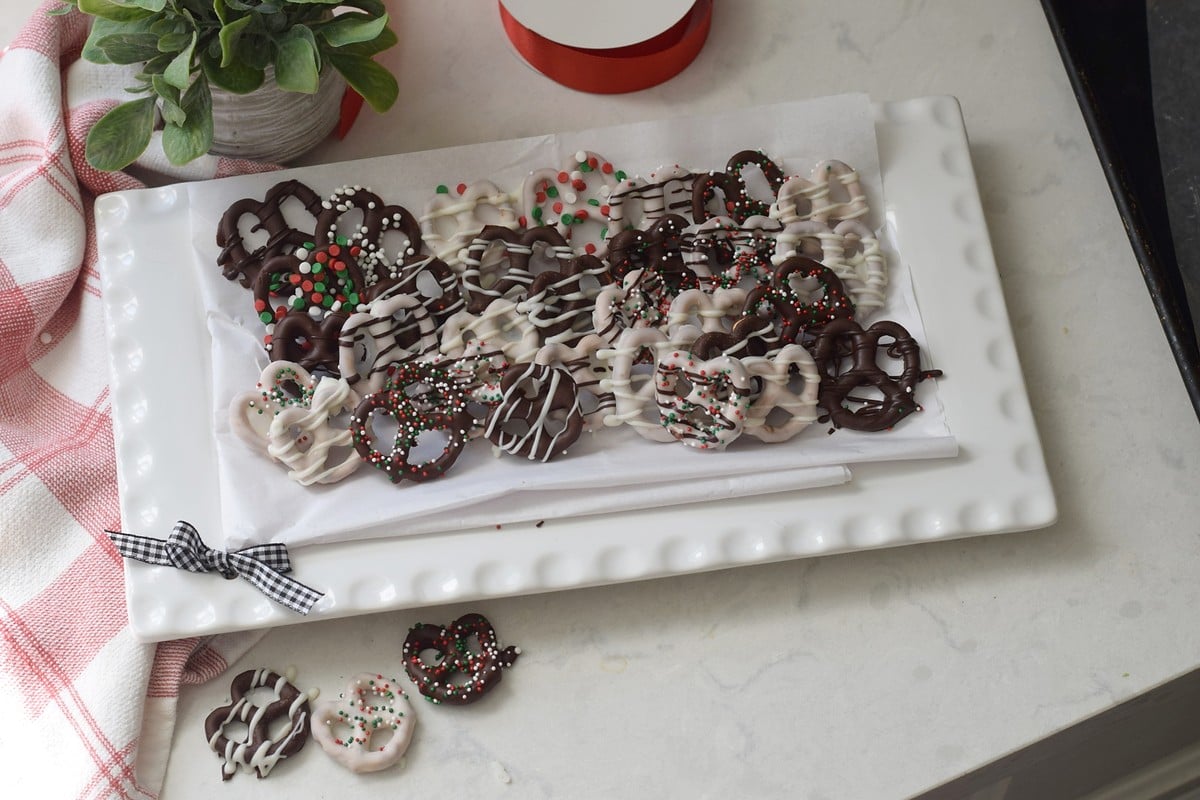 Platter of decorated chocolate covered pretzels beside a red-striped kitchen towel and potted plant.