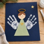 Handprint angel with white painted wings, a green paper dress, and a gold pipe cleaner halo on navy paper