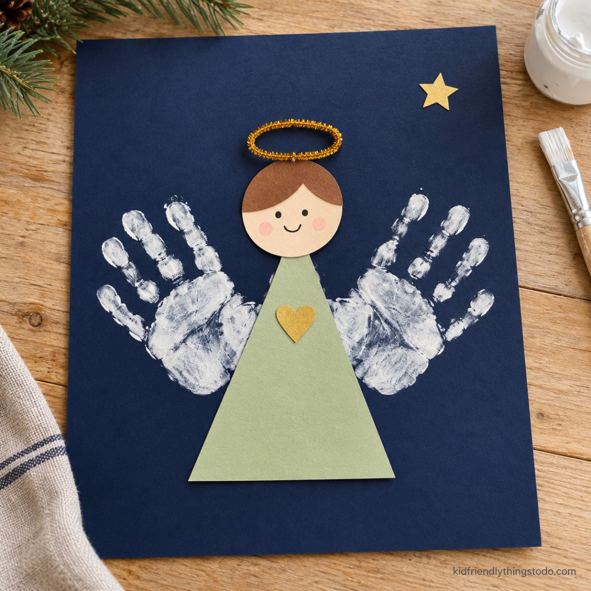 Handprint angel with white painted wings, a green paper dress, and a gold pipe cleaner halo on navy paper