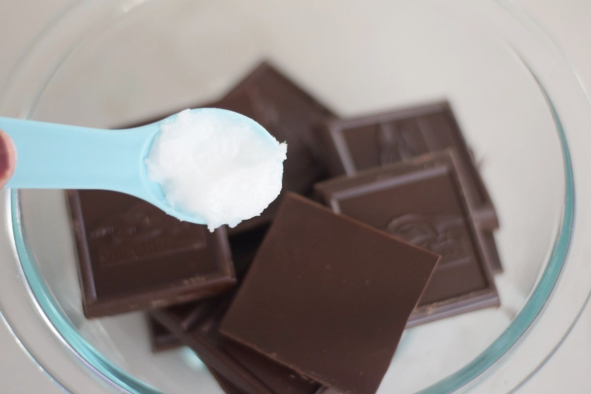 Adding coconut oil to broken dark chocolate baking bars in a glass bowl.