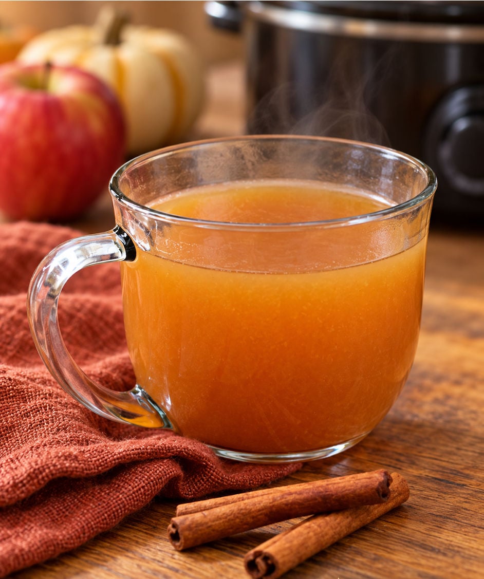 Warm mulled apple cider in a clear glass mug with cinnamon sticks on a wooden table and a slow cooker in the background.