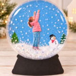 Snow globe bulletin board craft with a child's photo, snowy background, evergreen tree, and black base