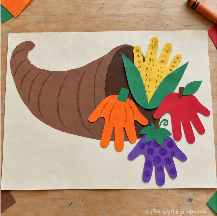 Handprint cornucopia craft for kids with colorful paper handprint fruits and vegetables spilling out of a Thanksgiving cornucopia.